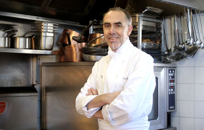 Bruno Seillery Chef Bruno Seillery souriant bras croisés dans une cuisine professionnelle avec des casseroles, poêles et ustensiles suspendus en arrière-plan