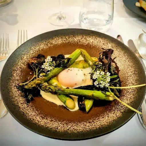 Restaurant l’Ambroisy plat 6 Assiette gastronomique avec asperges vertes, œuf poché, purée blanche et garniture de feuilles et fleurs comestibles sur une table avec couverts et verre en arrière-plan.